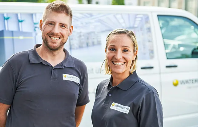 Ein Waterkotte Servicetechniker und eine Servicetechnikerin stehen in grauen Poloshirts mit Waterkotte Logo vor Servicewagen und lächeln. 