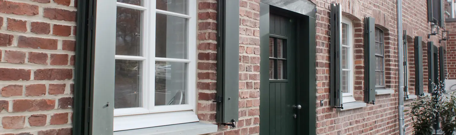 Close-up detail of a brick facade featuring a green entrance door and windows with dark green shutters.