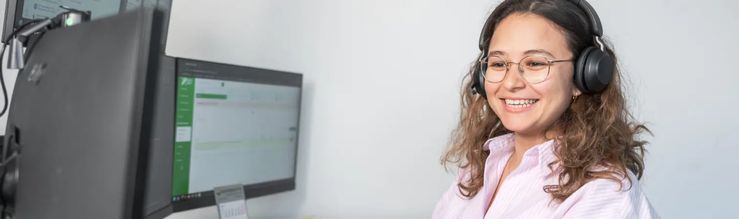 A service employee wearing a headset smiles, talks on the phone, and works on the computer.