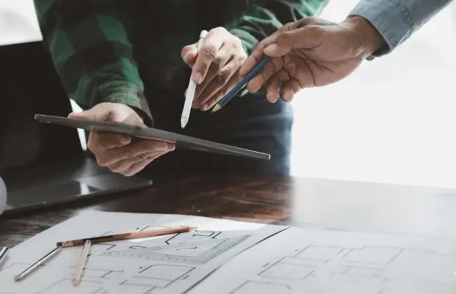 Hands of two people standing at a table with a laptop and construction plans; one person is holding a tablet.
