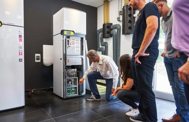 The instructor explains the training topics to the participants using an open heat pump in the practical training area of the Waterkotte Academy.