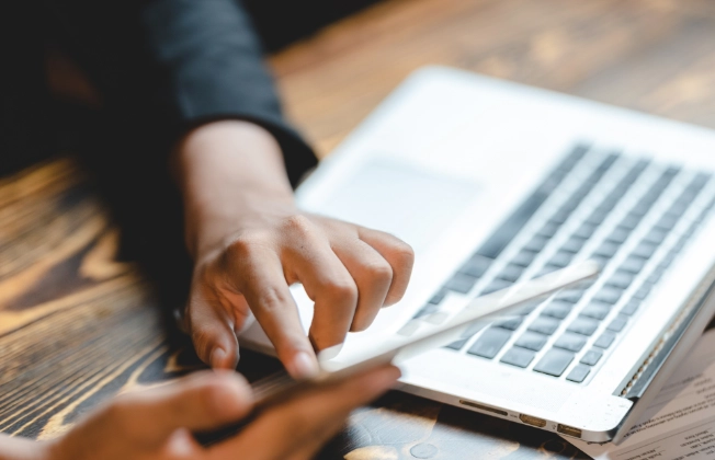 Close-Up auf Hände einer Person, die Tablet hält. Ein Laptop steht auf dem Tisch.