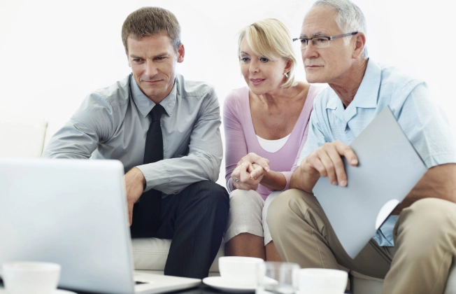 A man wearing a shirt explains something on a laptop to a couple standing in front of him.