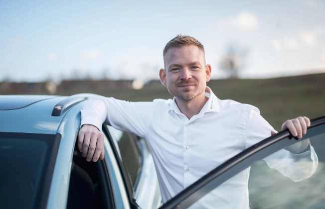 Smiling sales representative in a shirt leaning against an open car door. 