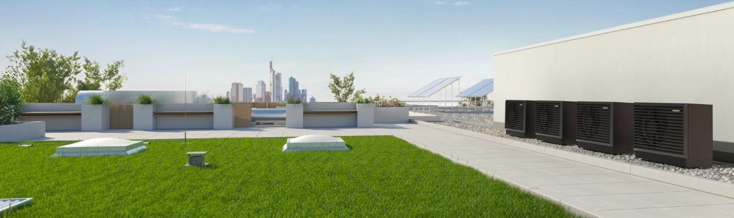 Vier Dunkelgraue Luftwärmepumpen-Außeneinheiten in Kaskade auf dem begrünten Dach eines modernen Gebäudes.